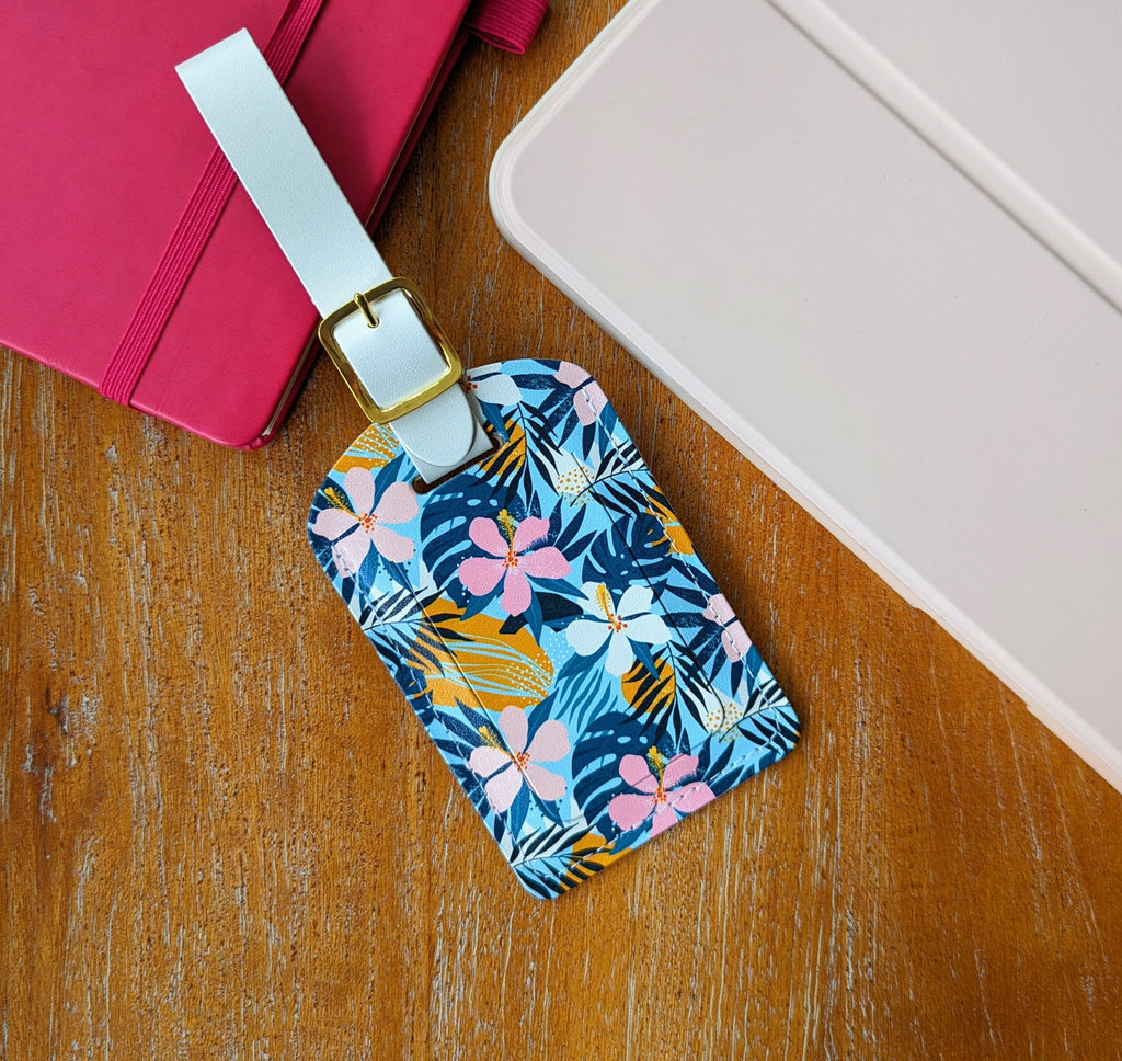 Recycled Leather Luggage Tag featuring tropical floral print, with flap covering personal details card, with a strap finished with a sleek gold-plated buckle to attach to luggage, partly on a pink notebook, next to a light pink ipad, on a wooden surface.