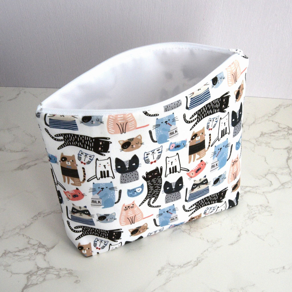 Large and generously sized cat print design wash bag with a 100% cotton outer, open, standing at an angle, with a white waterproof PU coated polyester lining, fastening with a quality white nylon zip, and a gusseted boxed bottom, so that it sits upright, placed on a marble surface, with a white background.