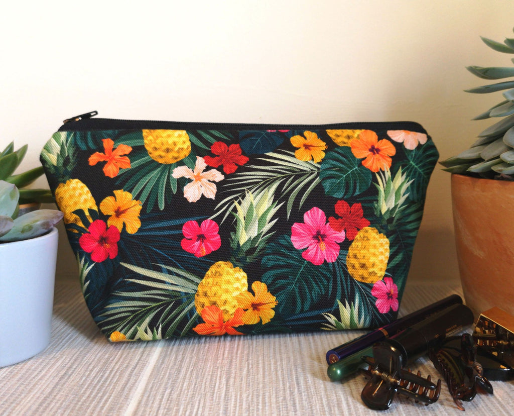 Tropical Makeup Bag featuring tropical pineapple floral print, with a black zip, sitting upright and closed, between two pot plants, next to some make up and two claw clips, on a bathroom shelf.