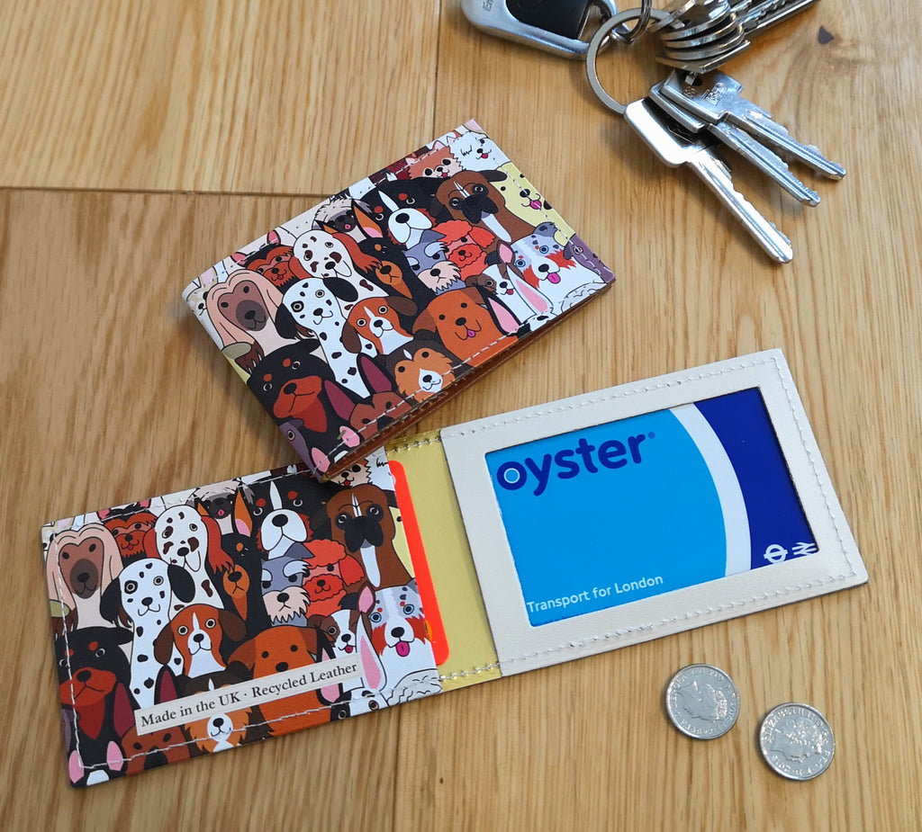 Recycled Leather Travel Card Holder featuring cute dog print, with oyster card in, lying open flat on wood, next to another closed travel card holder, placed next to a set of keys and coins to show scale.