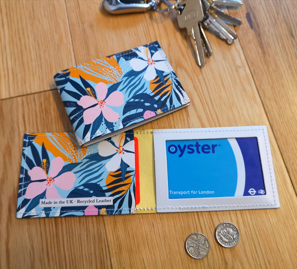 Recycled Leather Travel Card Holder featuring a tropical floral print, with oyster card in, lying open flat on wood, next to another closed travel card holder, placed next to a set of keys and coins to show scale.