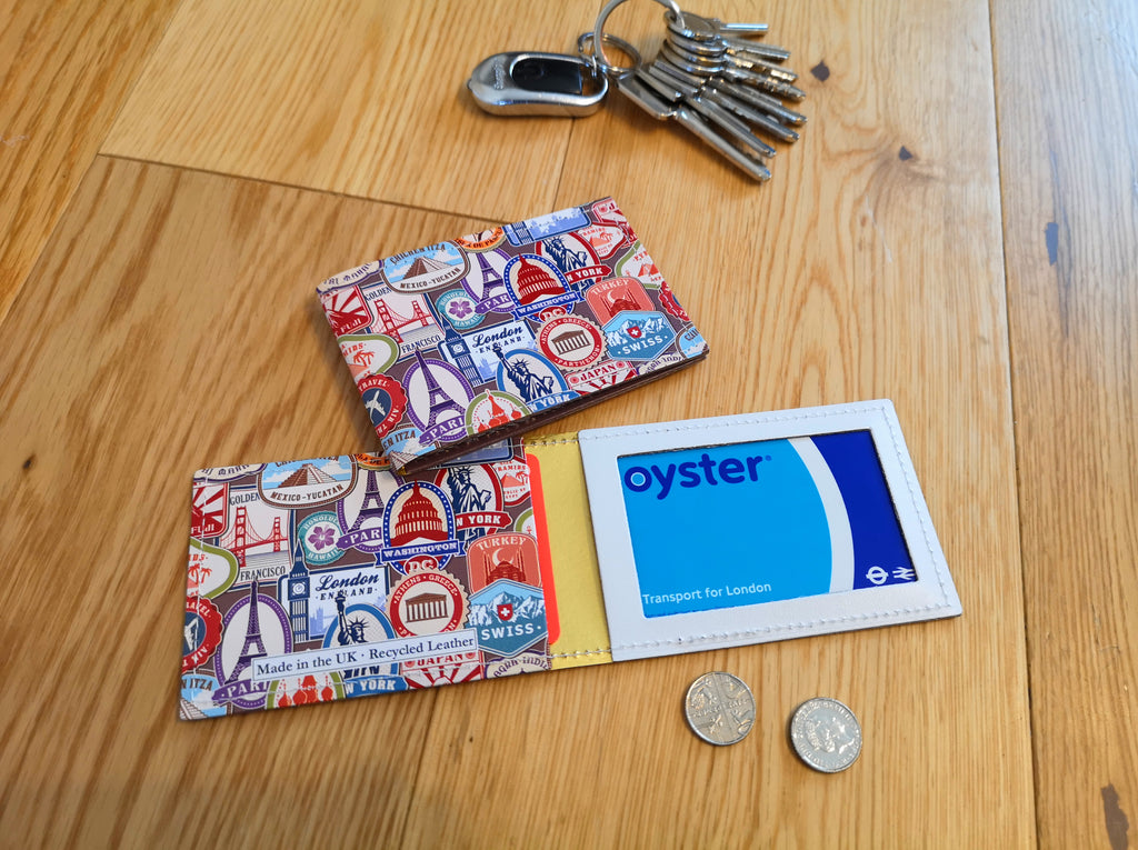 Recycled Leather Travel Card Holder featuring travel themed print, with oyster card in, lying open flat on wood, next to another closed travel card holder, placed next to a set of keys and coins, to show scale.