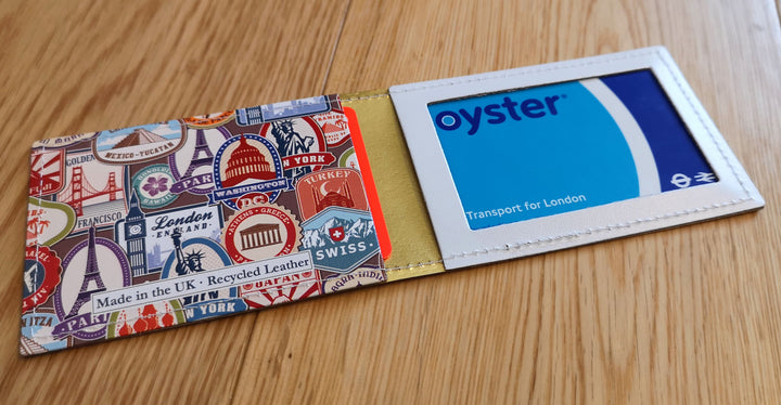 A travel themed travel card holder, with an oyster card in the window slot and a debit card in the other multi-purpose pocket, lying open flat on a wooden background.