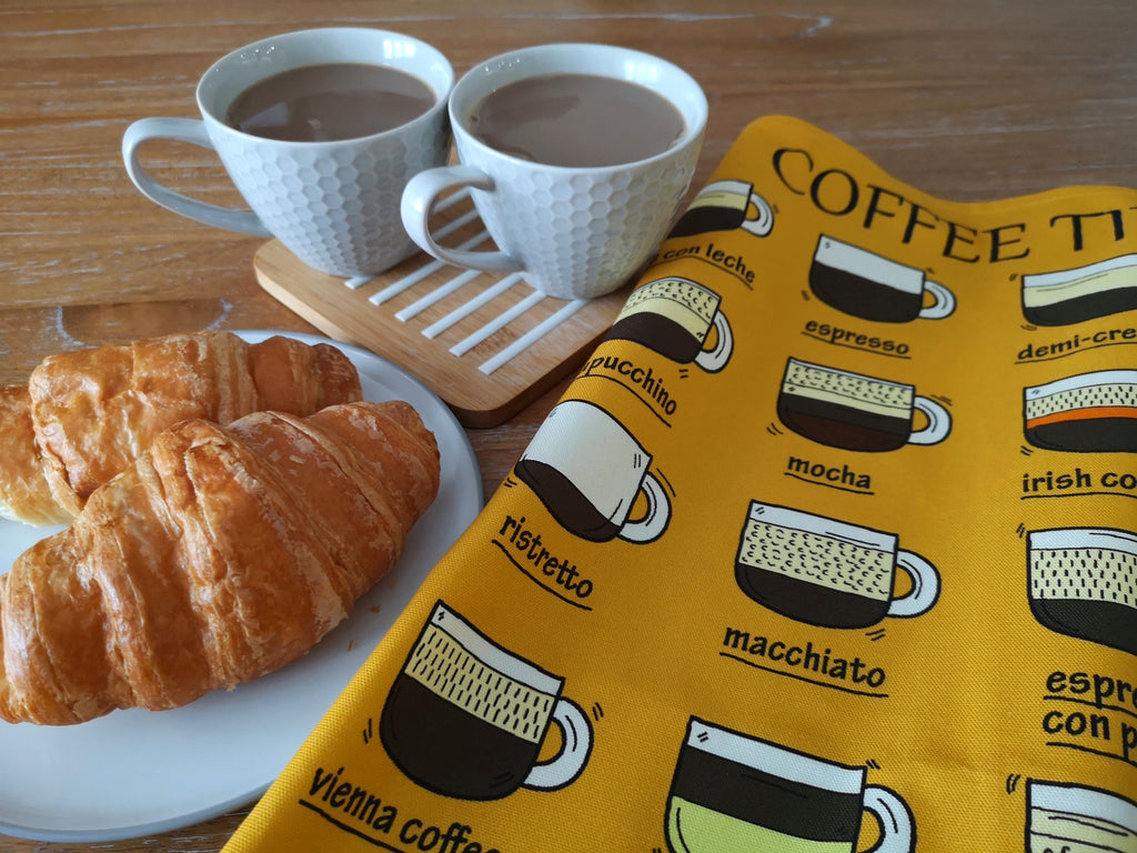 Coffee Themed Yellow Organic Cotton Tea Towel, next to two cups of coffee and two croissants on a white plate, all on a wooden table.