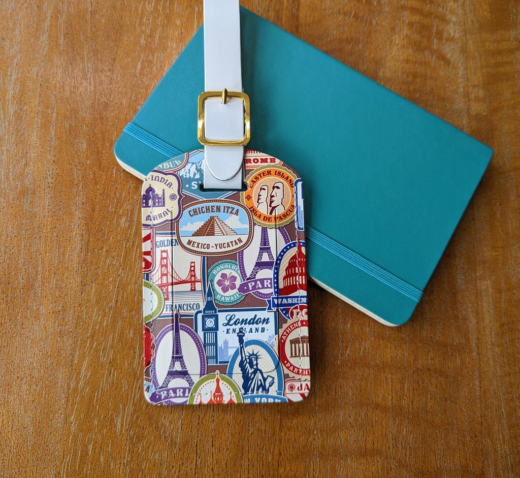 Recycled Leather Luggage Tag featuring travel themed print, with flap covering personal details card, with a strap finished with a sleek gold-plated buckle to attach to luggage, partly on a blue notebook, on a wooden surface.