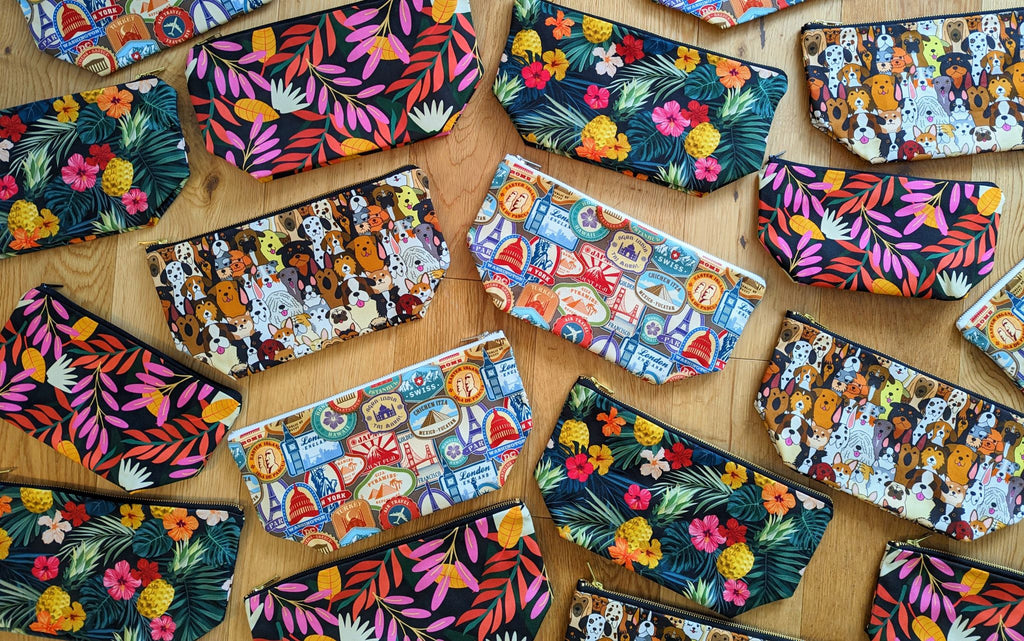 A large selection of wash bags, makeup bags, large washbags, large toiletry bags and zip pouches for travel in many different colours and patterns, including travel stamps, dogs, floral and pineapple designs, on a wooden background.