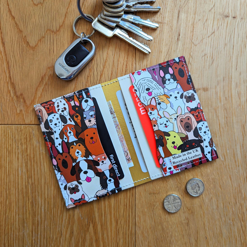 Recycled Leather Folding Wallet Card Holder featuring dog themed print, with four slots with cards and cash in, lying flat, at an angle, next to keys and coins, on a wooden surface.