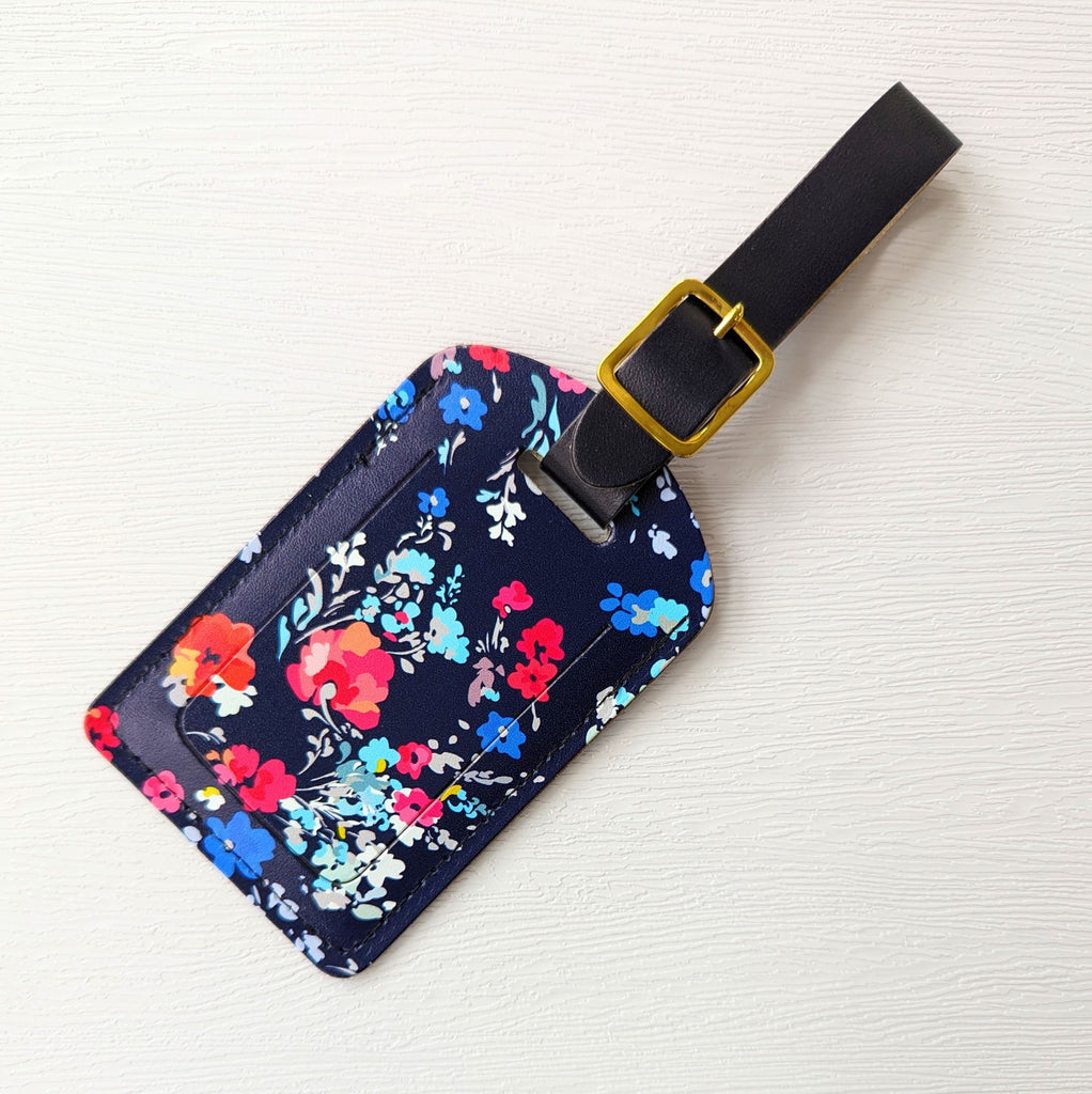 Recycled Leather Luggage Tag featuring dark blue floral print, with flap covering personal details card, with a strap finished with a sleek gold-plated buckle to attach to luggage, on a white background.