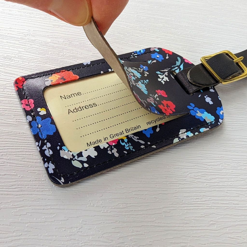 Recycled Leather Luggage Tag featuring dark blue floral print pattern, with strap finished with a sleek gold-plated buckle, to attach to luggage, close up, with privacy flap held open to reveal the personal details card, on a white background.