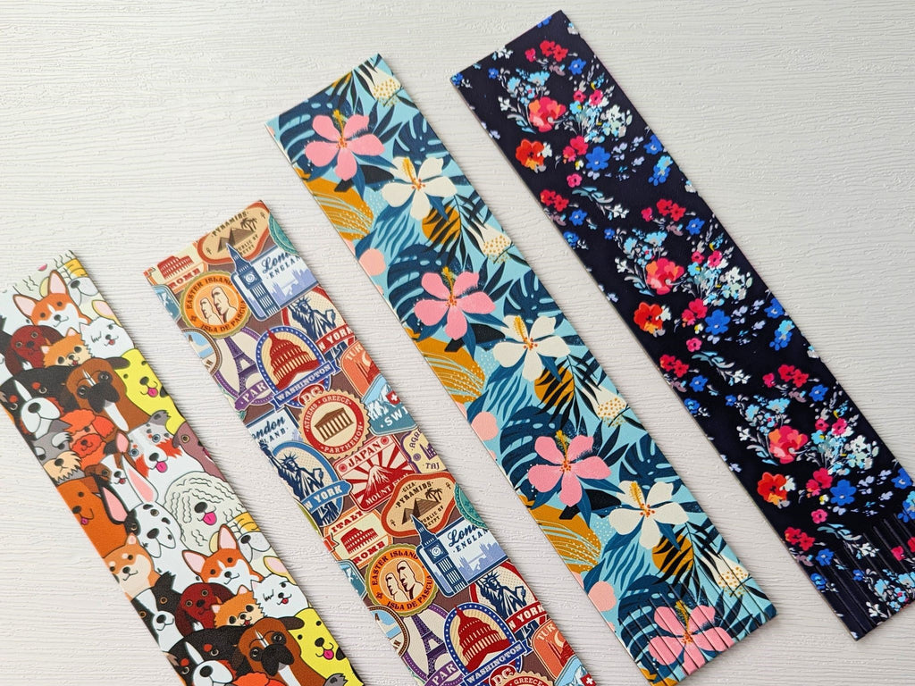 Four fringed recycled leather patterned bookmarks, one with a dog pattern, one with a travel stamps pattern, one with a tropical flower pattern and the other with a classic floral pattern on a dark blue background, all lying on a white background.