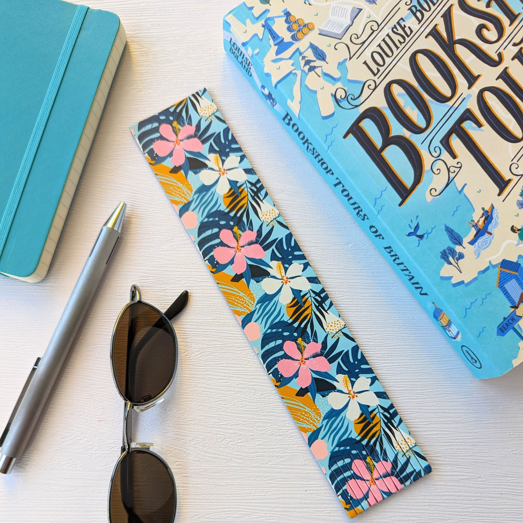 Bookmark, a recycled leather bookmark featuring a tropical floral print, lying on a white surface, next to a book, a pen, sunglasses, and a notebook. The colourful, vibrant floral bookmark is elegantly fringed at the bottom.