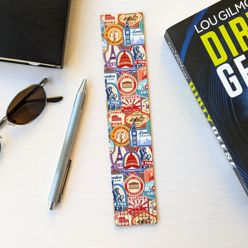 Bookmark, a recycled leather bookmark featuring a travel stamps pattern, lying on a white background, next to a book, a pen, some sunglasses, and a notebook. The travel bookmark is elegantly fringed at the bottom.