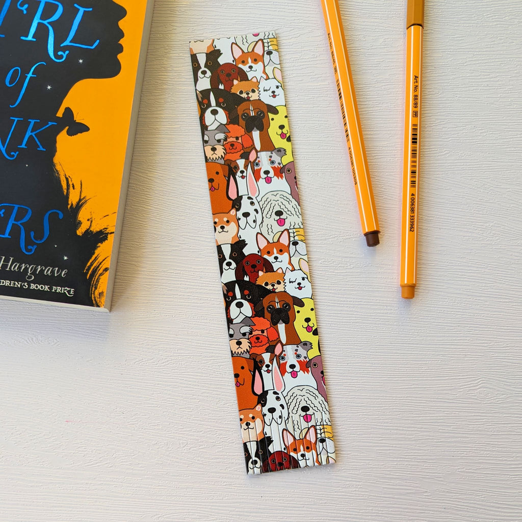 Bookmark, a recycled leather bookmark featuring a colourful dog print pattern lies on a white surface, next to a book and two colouring pens. The handmade dog bookmark is elegantly fringed at the bottom.