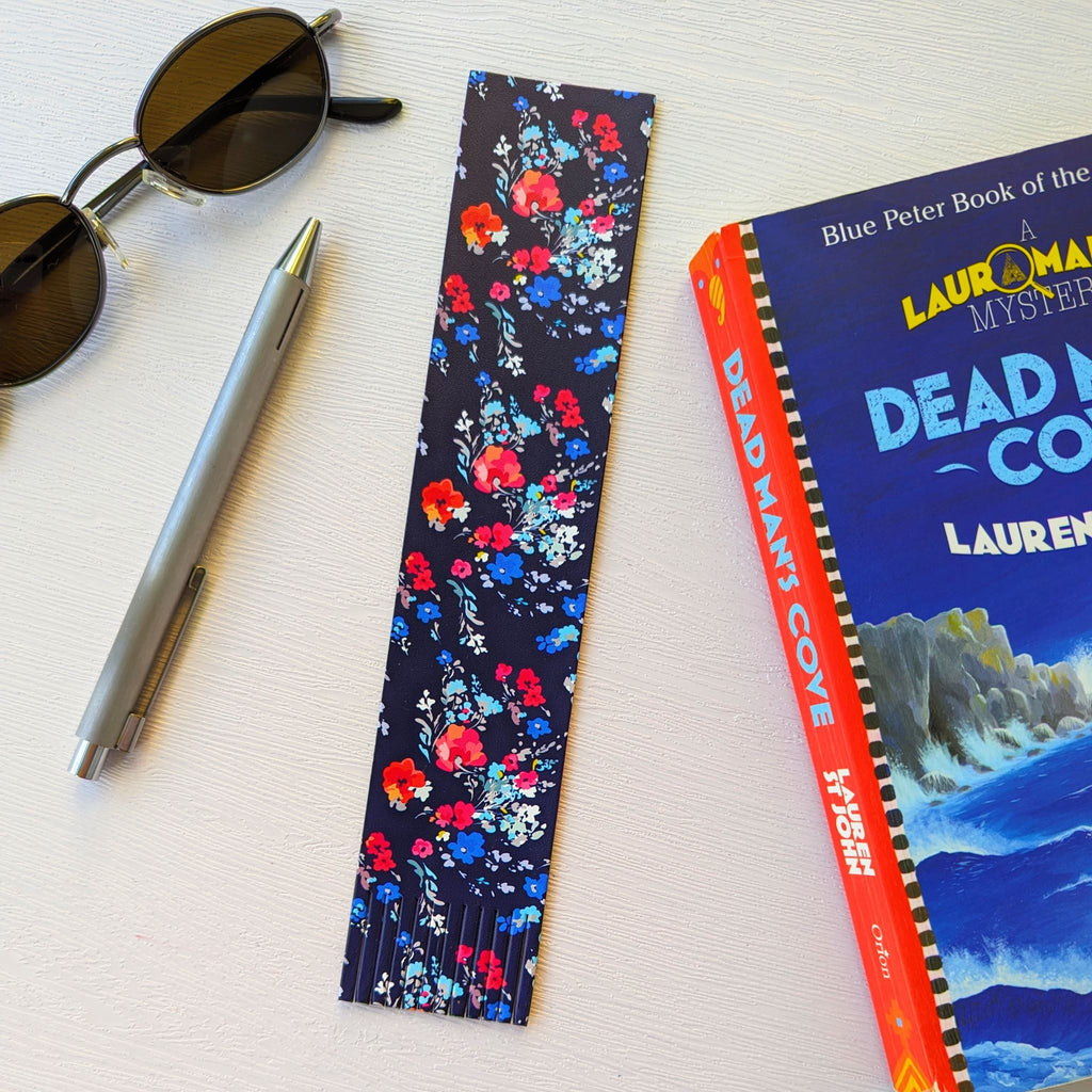 Bookmark, a recycled leather bookmark, featuring a floral print on a dark blue background, the bookmark is lying on a white surface, next to a book, a pen, and sunglasses. The floral bookmark is elegantly fringed at the bottom.