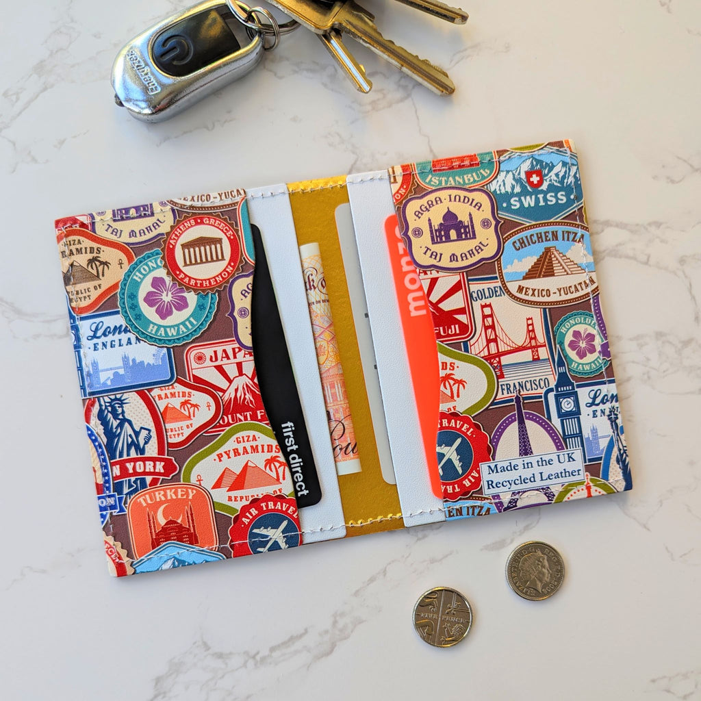 Recycled Leather Folding Wallet Card Holder featuring travel themed print, with four slots, with cards and cash in, lying open and flat, next to keys and coins on a wooden surface.