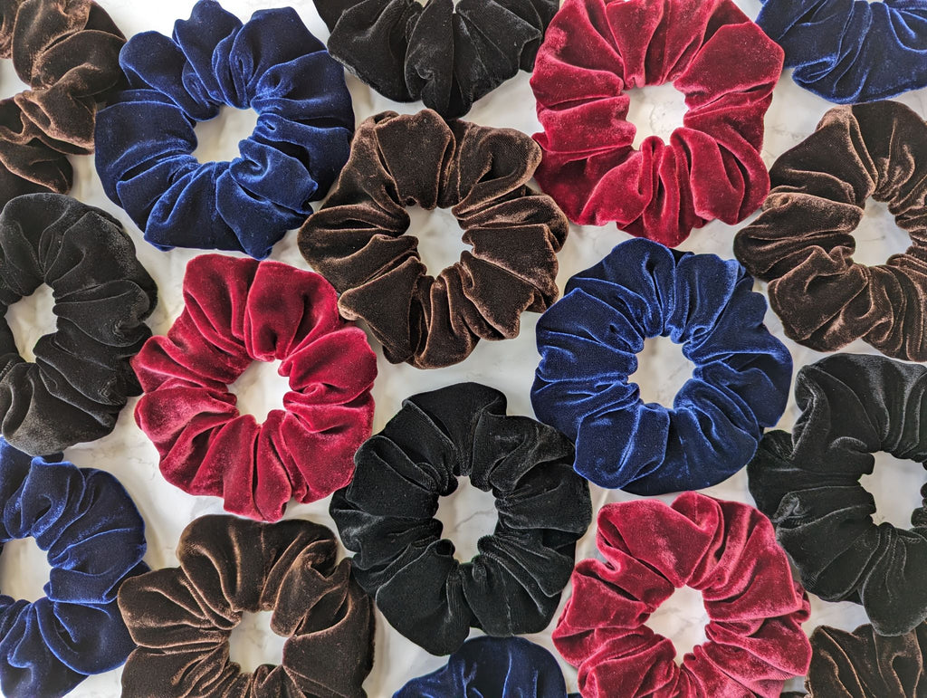 A large collection of luxurious silk velvet hair scrunchies, in black, navy blue, dark red and dark brown, on a plain marble background.