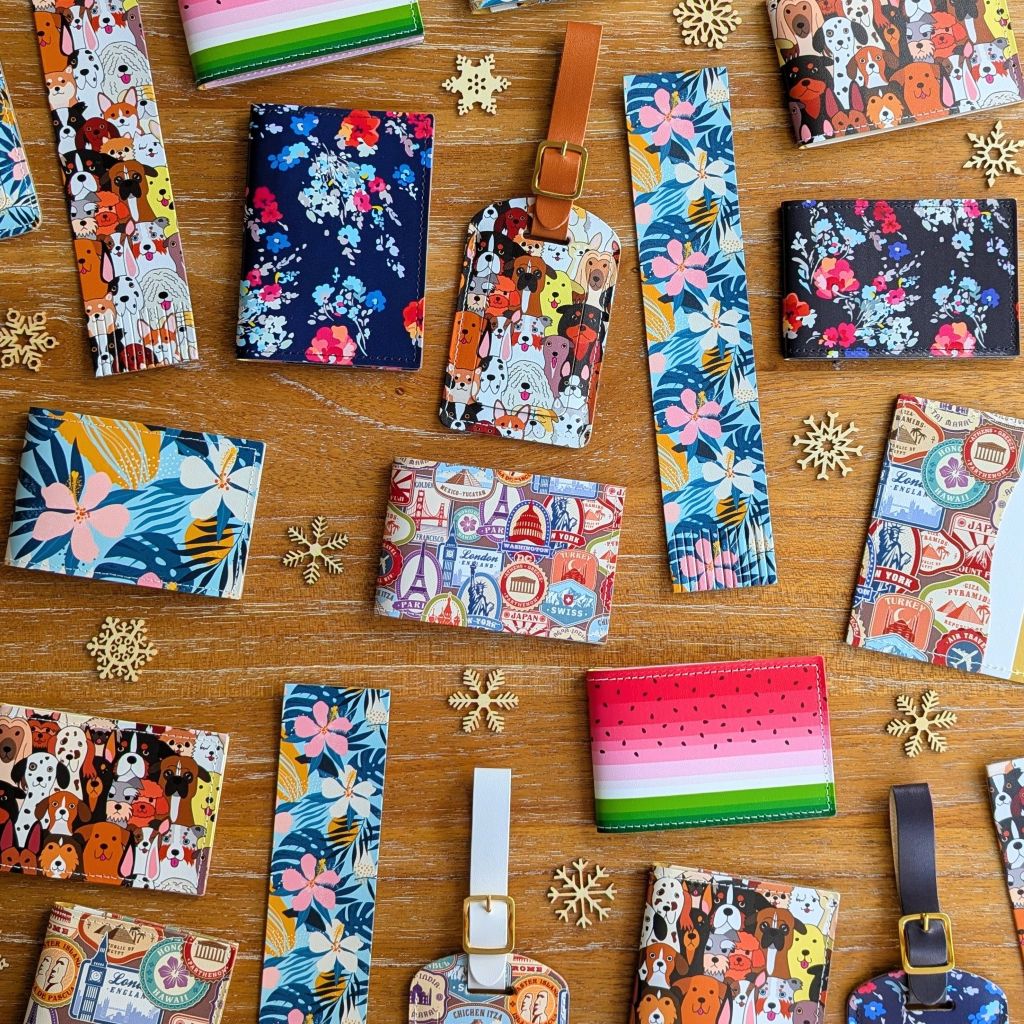 A large collection of colourful and vibrant recycled leather products, including travel card holders, oyster card holders, folding wallet card holders and luggage tags, with a range of patterns including dogs, travel stamps, tropical flowers, and classic flowers on dark blue, all on a wooden surface, with small decorative snowflakes. 