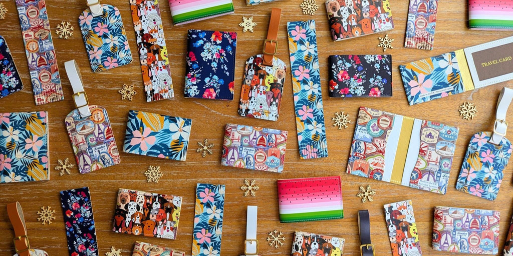 A large collection of colourful and vibrant recycled leather products, including travel card holders, oyster card holders, folding wallet card holders and luggage tags, with a range of patterns including dogs, travel stamps, tropical flowers, and classic flowers on dark blue, all on a wooden surface, with small decorative snowflakes. 