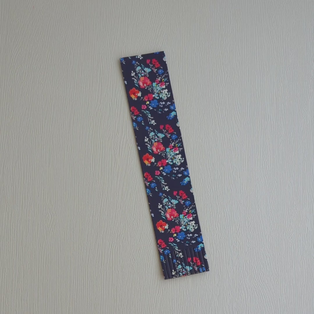 A recycled leather bookmark, featuring a floral print on a dark blue background, lying on a white surface, is picked up and turned over and then placed inside a book.