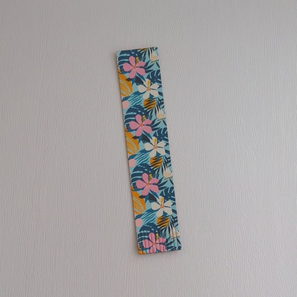 A recycled leather fringed bookmark featuring a tropical floral print, lying on a white surface, picked up and turned over and then placed in a book.