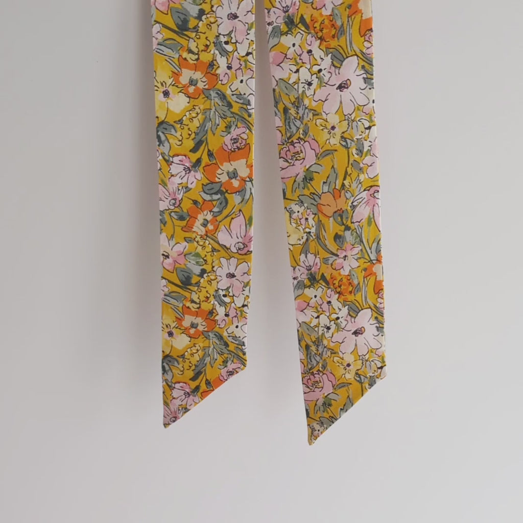 Luxurious silk satin skinny scarf, crafted from 100% pure silk satin and adorned with a stunning floral design, featuring delicate pink and orange flowers on a vibrant mustard yellow background, hanging on a white background, lifted to show the details.