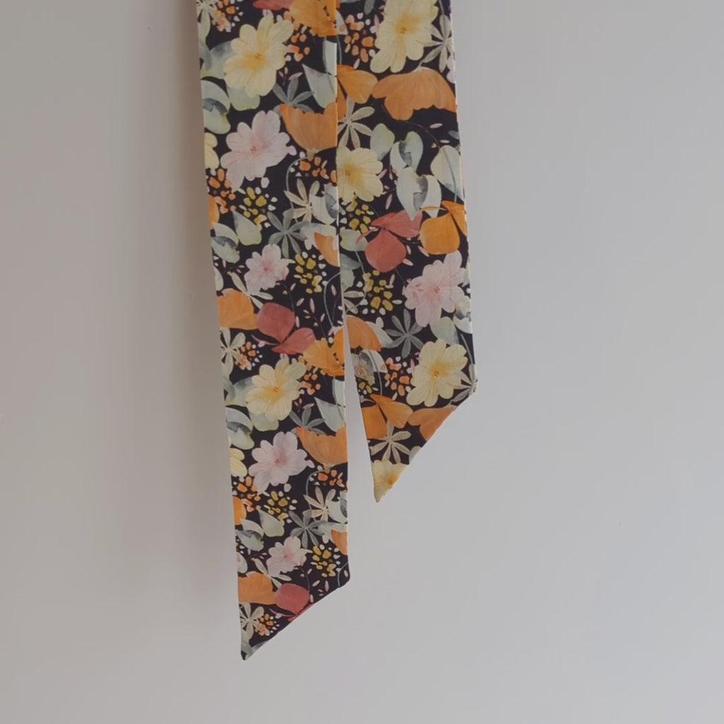 Luxurious silk satin skinny scarf, crafted from 100% pure silk satin and adorned with a stunning floral design, featuring orange and pink flowers on a black background, hanging on a white background, slowly lifted to show the detail.