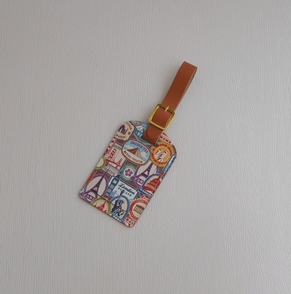 Recycled Leather Luggage Tag featuring travel themed print pattern of famous landmarks, with flap covering personal details card, with a brown strap finished with a sleek gold-plated buckle to attach to luggage, lying on a white background, picked up and turned over, then the flap opened to reveal the personal details card inside.