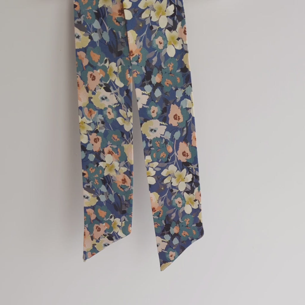 Luxurious silk satin skinny scarf, crafted from 100% pure silk satin and adorned with a stunning floral design, featuring pink, white and teal flowers on a blue background, hanging on a white background and lifted to show the details.