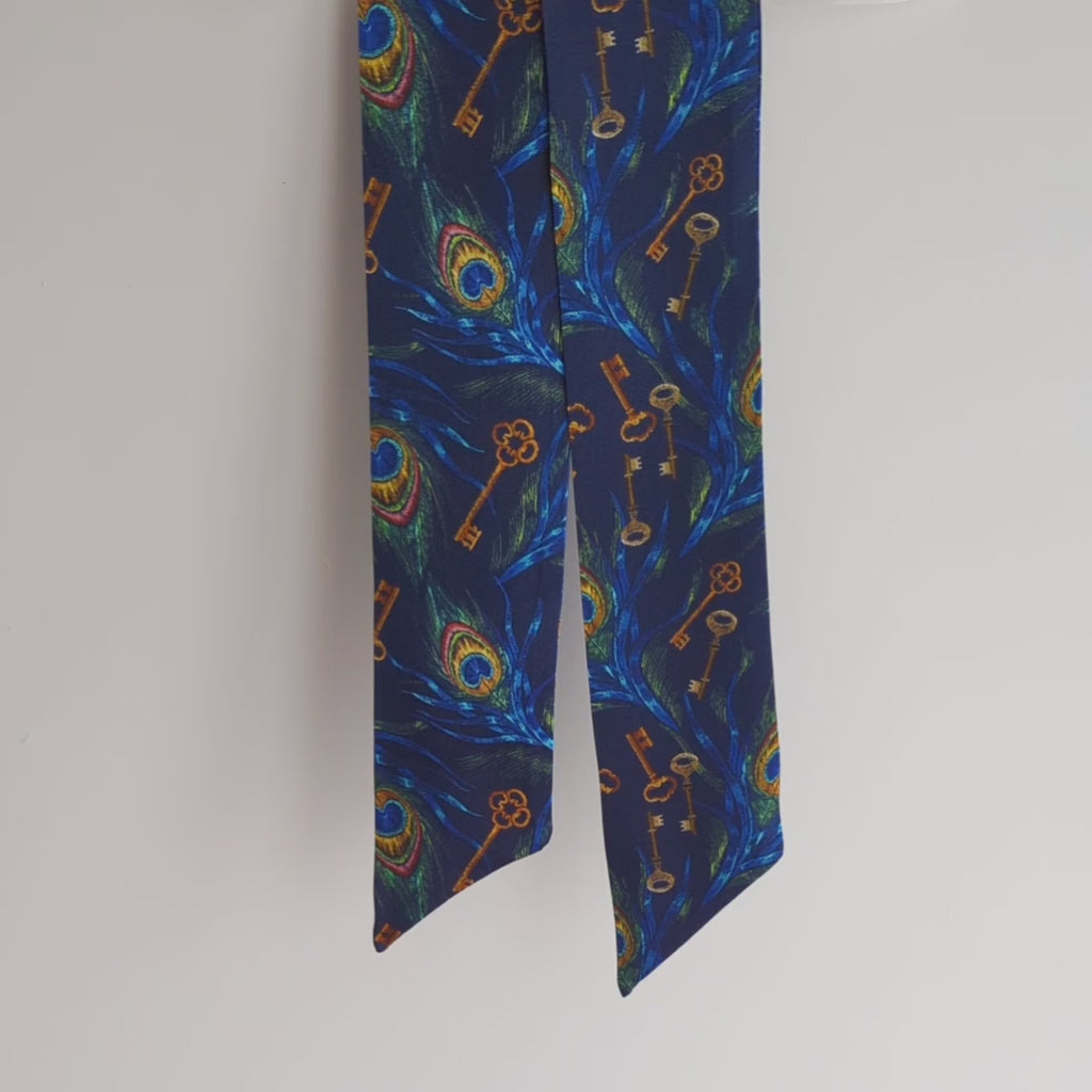 Luxurious dark blue silk satin skinny scarf, crafted from 100% pure silk satin and adorned with a stunning design, featuring peacock feathers and keys, hanging on a white background, lifted to show the details.