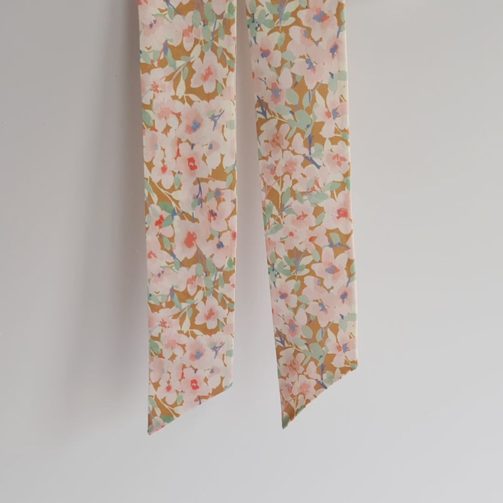Luxurious silk satin skinny scarf, crafted from 100% pure silk satin and adorned with a stunning pink cherry blossom design, on a light beige background, hanging on a white background and lifted to show the details.